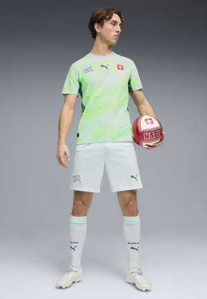Joven futbolista masculino con uniforme verde y blanco del equipo suizo sosteniendo un balón de fútbol rojo y blanco con la cruz suiza.