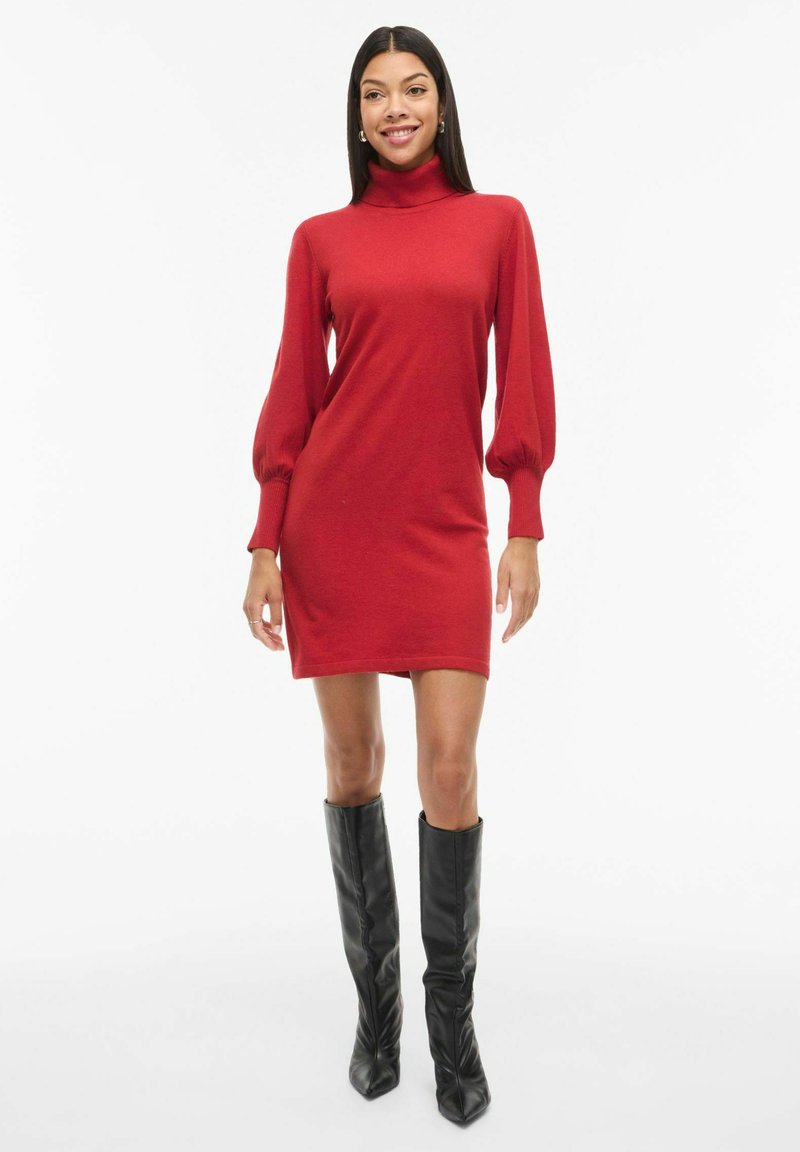 Vestido rojo de manga larga con cuello alto, que presenta una silueta ajustada y tela texturizada. Lleva botas negras hasta la rodilla.