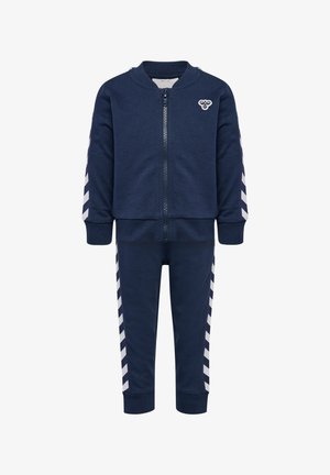 Granatowy dres zapinany na zamek z okrągłym kołnierzem, z białymi paskami w kształcie chevronu na rękawach i spodniach. Wykonany z teksturowanej tkaniny.