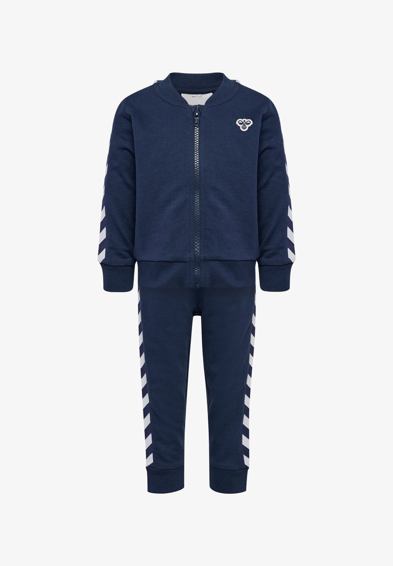 Marinblå zip-up träningsoverall med rund krage, med vita chevronränder längs ärmarna och byxorna. Tillverkad av texturerat tyg.