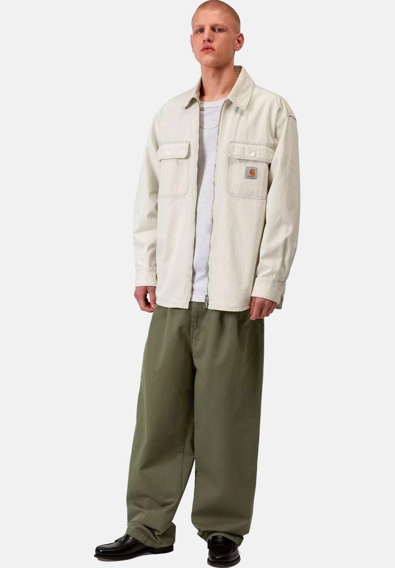 Hombre de pie con una chaqueta de pana crema sobre una camisa blanca, pantalones verdes oliva holgados y zapatos de cuero negros sobre un fondo liso.