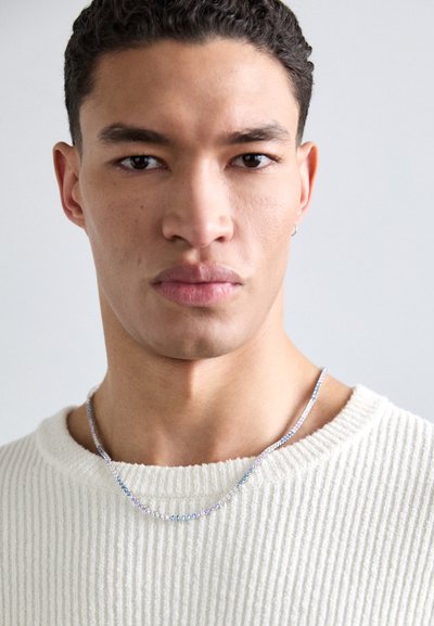 Collar de cadena de plata con pequeños acentos multicolores. Lleva puesto sobre un suéter blanco texturizado. Primer plano del rostro de la persona que lo lleva.
