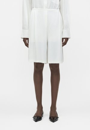 Μοντέλο που φορά λευκά tailored σορτς μέχρι το γόνατο, συνδυασμένα με λευκό πουκάμισο με μακριά μανίκια και μαύρα μυτερά φλατ παπούτσια, στέκεται μπροστά σε λευκό φόντο.