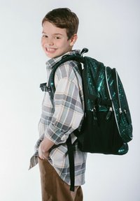 Lo zaino presenta una base nera con accenti in un luminoso motivo digitale verde, spallacci imbottiti e numerosi scomparti. Indossato da un ragazzo in una camicia a quadri.