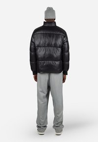 Mann iført svart blank pufferjakke, lysegrå vide bukser, grå strikket lue og hvite sneakers, står med ryggen til mot hvit bakgrunn.