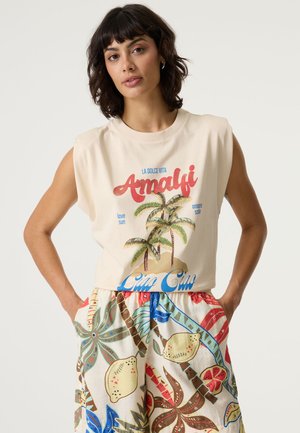 Haut sans manches crème avec graphismes de palmiers et texte rouge et bleu, assorti à une jupe multicolore avec motifs de feuilles tropicales et de fruits.