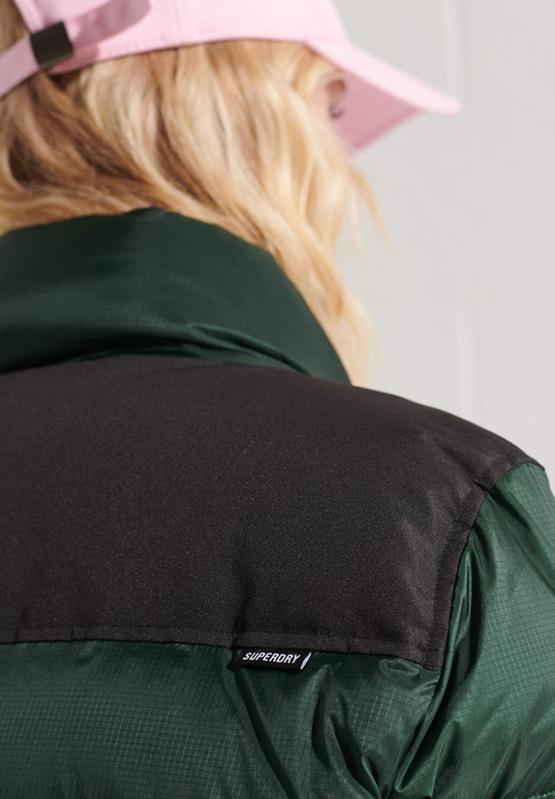 Czarna i ciemnozielona pikowana kurtka z kołnierzem o gładkiej fakturze, z widoczną metką marki i różową czapką w tle.