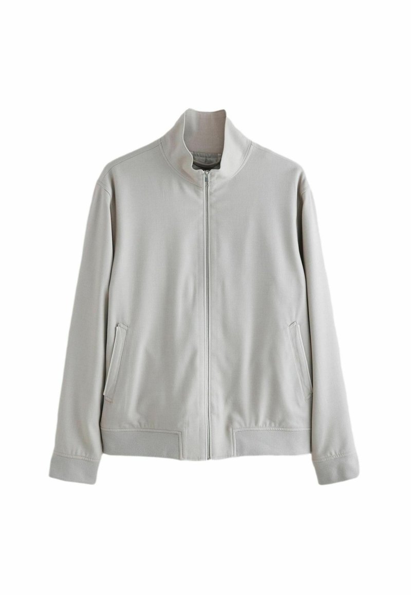 Gai pelēks rāvējslēdzēja mētelis, izgatavots no gludas auduma, ar augstu apkakli, divām sānu kabatām un ribotām aprocēm un apakšmalu.