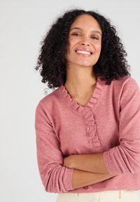 Maglione rosa con scollatura a V, colletto arricciato e dettagli con bottoni. Realizzato in materiale morbido con maniche a coste. Design casual.