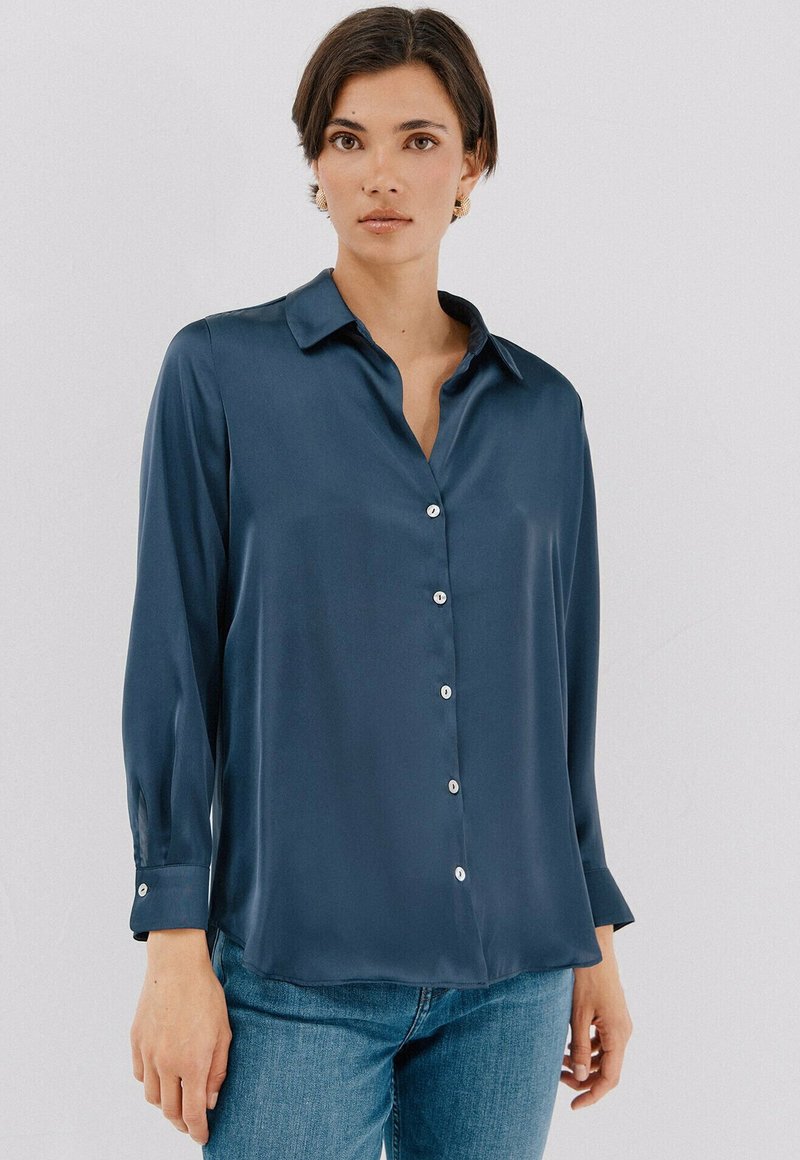 Blusa di raso blu navy con colletto, maniche lunghe e chiusura con bottoni. Presenta una sottile lucentezza e polsini con bottoni, abbinata a denim casual.