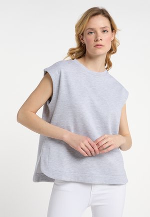 Ung kvinde med lyst krøllet hår iført en ærmeløs lysegrå top og hvide bukser, poserende med hænderne foldet foran.