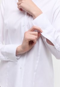 White button-up shirt with a textured fabric; features a rounded collar and buttoned cuffs, showcasing a hand fastening a button.