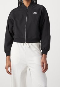 Chaqueta bomber corta negra con logo blanco de Puma, cierre de cremallera y puños acanalados. Tejido liso con un corte ligeramente oversize.
