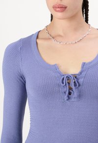 Texturált anyagból készült, hosszú ujjú, lila blúz, amelynek kerek nyakkivágása fűzős részlettel és testhezálló sziluettel rendelkezik.
