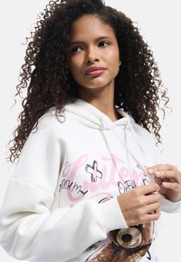 Witte hoodie met een grafisch ontwerp van een beer en speelse tekst. De stof lijkt zacht te zijn; het ontwerp heeft roze en zwarte accenten.