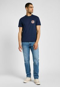 Mörkblå bomullströja med en röd och vit logotyp på vänster bröst, i kombination med ljusblå denimjeans och krämfärgade sneakers.