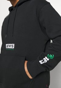 Sudadera negra con capucha, bolsillo delantero y cordones, con el logo de Levi's impreso en blanco y verde en el pecho y la manga. Textura suave.