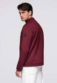 Bordeauxfarbige Jacke mit strukturierter Oberfläche, hohem Kragen und schwarzem Akzent-Patch auf der linken Schulter. Weiße Hose ergänzt das Design.