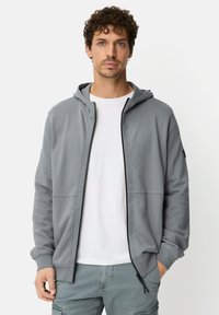 Grijze zip-up hoodie gemaakt van zacht materiaal, voorzien van een capuchon, voorzakken en een gladde textuur. Gedragen over een wit T-shirt.