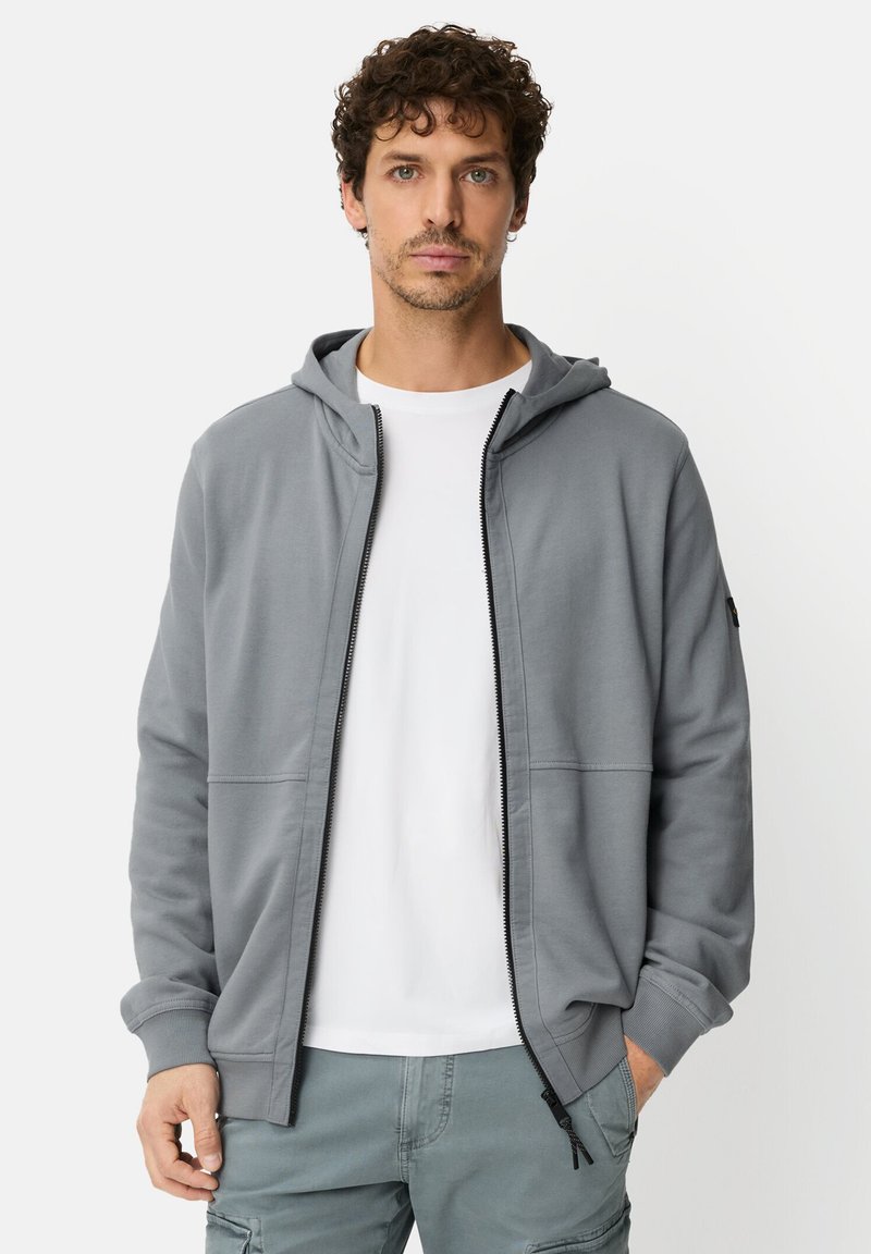 Grijze zip-up hoodie gemaakt van zacht materiaal, voorzien van een capuchon, voorzakken en een gladde textuur. Gedragen over een wit T-shirt.