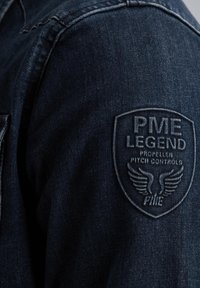 Granatowa kurtka dżinsowa o teksturowanej powierzchni, z naszywką z haftem przedstawiającą "PME LEGEND" oraz wzorami skrzydeł na ramieniu.