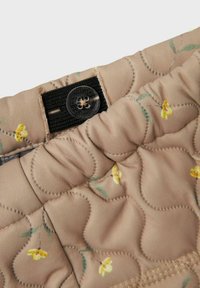 Quilted stof i blød beige med et blomstermønster, der har en sort velcro-strop og klare knapdetaljer.