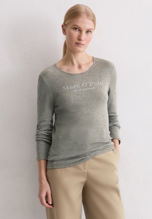 Długi, szary top z miękkiego materiału z okrągłym dekoltem. Z przodu znajduje się napis "Marc O'Polo" na wysokości klatki piersiowej. Połączony z jasnobeżowymi spodniami.
