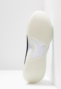 Czarna podeszwa sportowego buta z teksturowaną białą gumową podeszwą zewnętrzną, z okrągłymi uchwytami i gładkim, zakrzywionym akcentem w środkowej części.