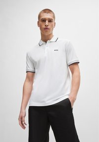Cămașă polo albă cu margine neagră la guler și manșete, având un logo mic. Modelul pozează casual, cu mâinile în buzunare, purtând pantaloni negri.