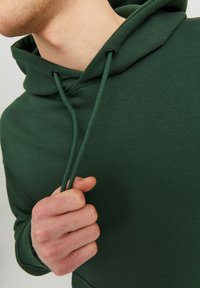 Felpa verde scuro con cordino in nylon, caratterizzata da una texture morbida e una vestibilità comoda. Dettaglio ravvicinato dell'area del collo e della mano che afferra il tessuto.