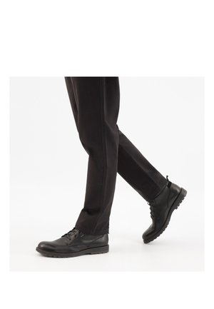 Person, die schwarze Schnürstiefeletten und schwarze Hosen trägt, geht mit einem leicht erhobenen Fuß vor einem weißen Hintergrund.