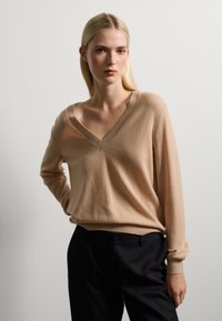 Suéter beige con cuello en V en suave tejido de punto, con mangas largas y puños acanalados, combinado con pantalones negros ajustados.
