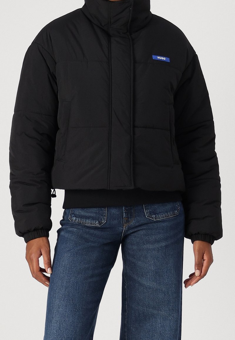 Personne portant une doudoune courte noire avec un logo Hugo bleu sur la poitrine et un jean en denim bleu sur un fond uni.