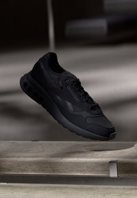 Sapatilha atlética preta com parte superior texturizada e sola perfurada, posicionada acima de uma superfície de concreto cinza claro em iluminação suave.