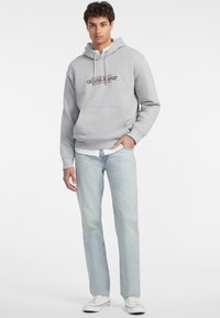 Felpa grigia con tasca frontale e coulisse, con logo "GUESS JEANS" in nero e rosso. Abbinata a jeans azzurri chiari e sneakers bianche.