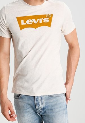 Mann trägt ein beiges Levi’s T-Shirt mit orangem Logo und hellblauen Jeans vor einfachem Hintergrund.