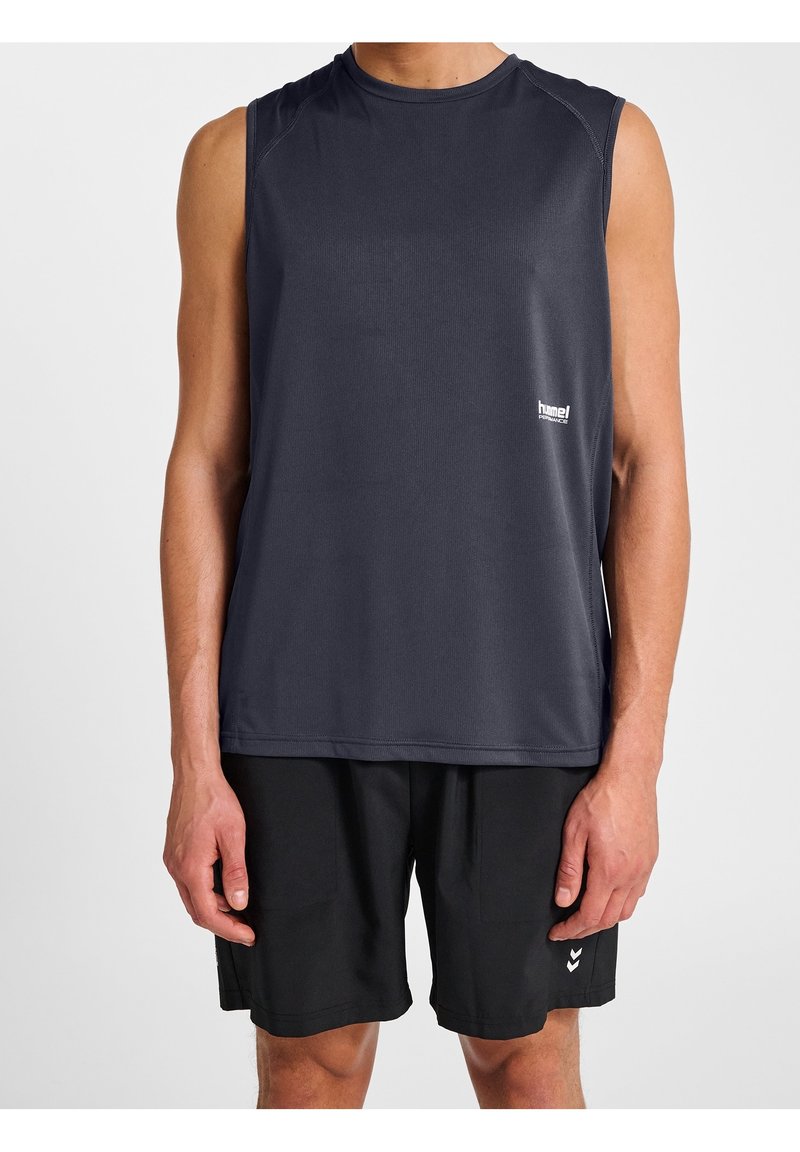 Homme portant un débardeur de sport gris foncé et un short noir, debout devant un fond blanc uni.
