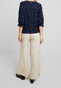 Námořnická blůza s vícbarenými vzory listů, nařasenými rukávy a hladkou texturou, zkombinovaná se béžovými širokými kalhotami a černými botami.