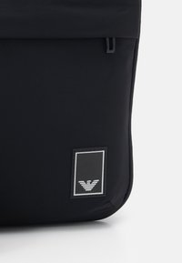 Svart ryggsäck gjord av slitstark tyg, med en rulltoppsdesign, slät yta och en silverlogotagg med ett randigt emblem.