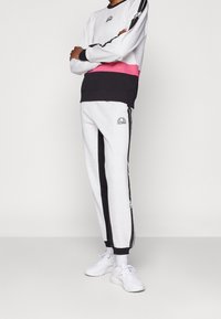 Sudadera gris con bandas horizontales negras y rosas, combinada con pantalones de chándal grises que presentan franjas laterales negras. Zapatillas blancas completan el look.
