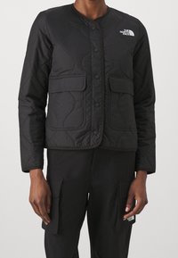 Černá prošívaná bunda s kulatým výstřihem, patentkami, dvěma předními kapsami a logem "The North Face" na levé horní straně.