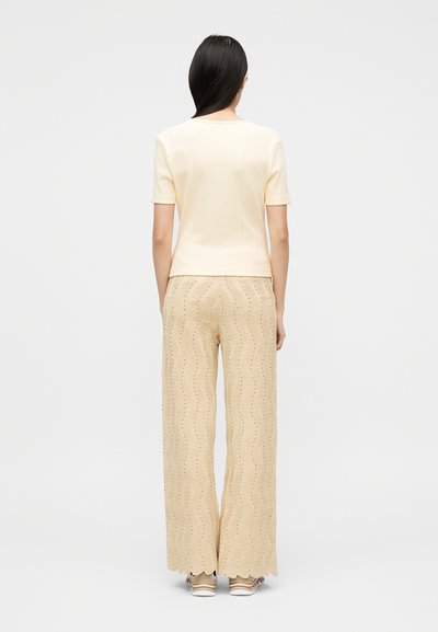 Γυναίκα με μακριά σκούρα μαλλιά, φοράει ένα κρεμ τοπ με κοντά μανίκια και μπεζ παντελόνι με φαρδιά μπατζάκια και σχέδια, στέκεται με την πλάτη στραμμένη στην κάμερα.