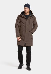 Ruskea vedenpitävä parkatakki, jossa on huppu, vetoketju- ja nepparikiinnitys sekä sivutaskut. Yhdistetty mustiin housuihin ja mustiin saappaisiin. Päällä sininen ribbineulottu pipo.