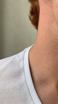 Vit bomullsskjorta med rund halsringning, som visar hud och ljus stubbe på halsen. Texturen verkar slät och mjuk.