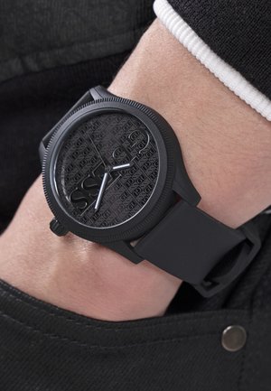 Mustast silikoonist rihmaga käekell, millel on tekstuurne Guessi logoga sihverplaat, kantud vasakul randmel musta riietuse üle.