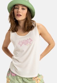Top bianco con design floreale rosa e testo dorato. Chi lo indossa indossa anche un cappello verde e pantaloni a motivi floreali.