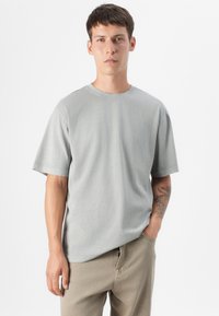 En ung mand står mod en lys baggrund, iført en grå T-shirt og beige bukser, med en tatovering synlig på sin venstre arm.