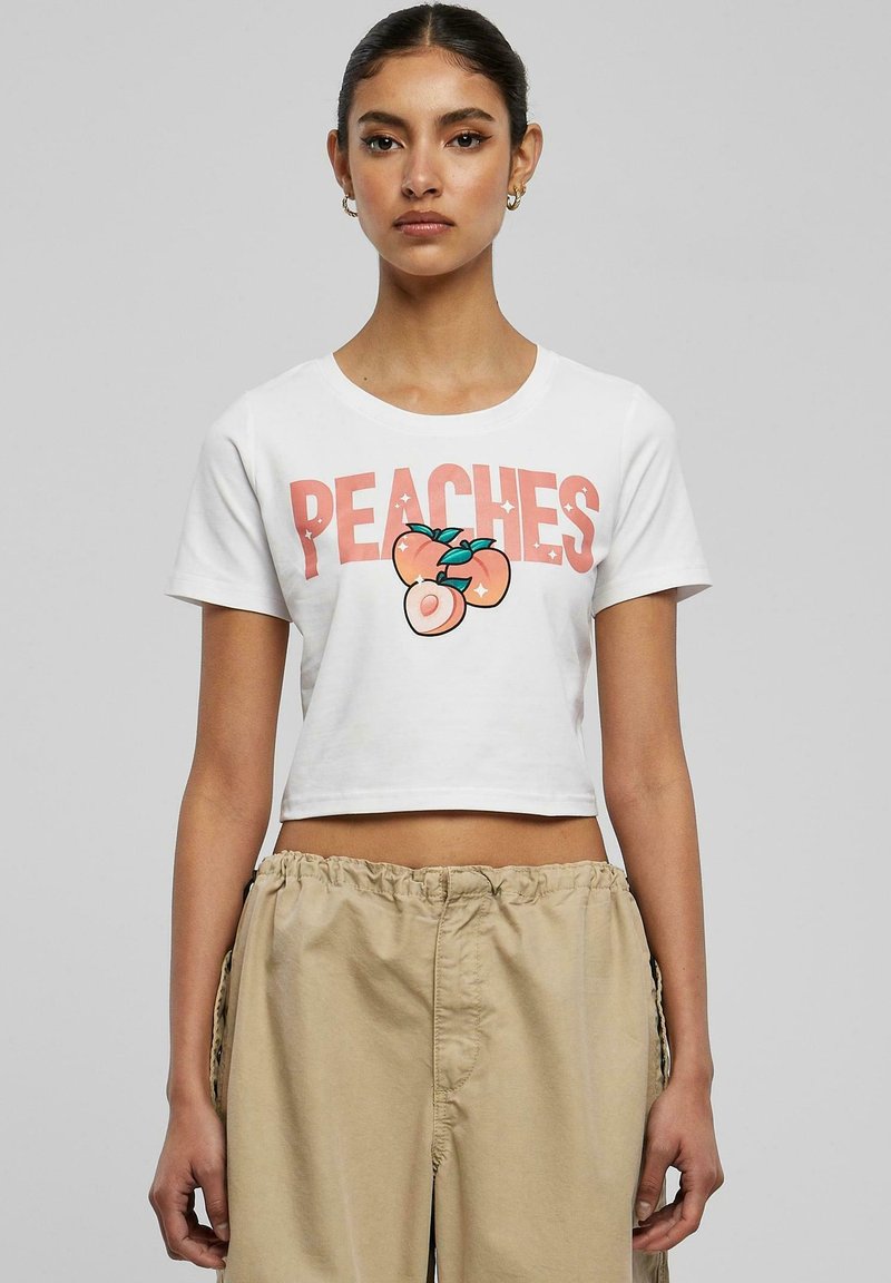 Balta īsā t-kreklā ar vārdu "PEACHES" rozā krāsā un ilustrāciju ar persikiem, apvienota ar bēšiem aukliņu biksēm.