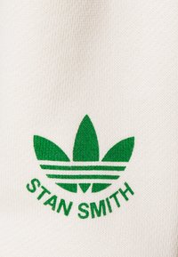 Zelené logo Adidas trefoil s textem "STAN SMITH" pod ním na bílém texturovaném pozadí.