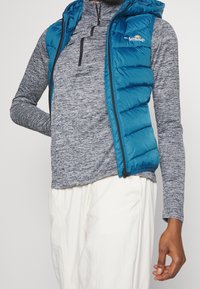 Blaues Steppweste mit Kapuze, gestepptes Texturmuster, kombiniert mit einem grauen Langarmshirt mit einem Viertelreißverschluss und einem dezenten Melangemuster.